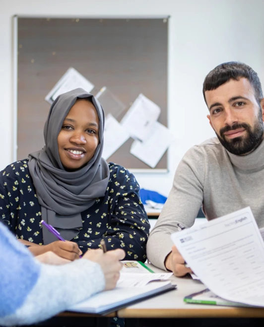 A group of students, including a male student in a gray turtleneck sweater and a female student in a gray hijab, engage in a discussion while reviewing documents together. A group of students, including a male student in a gray turtleneck sweater and a female student in a gray hijab, engage in a discussion while reviewing documents together.